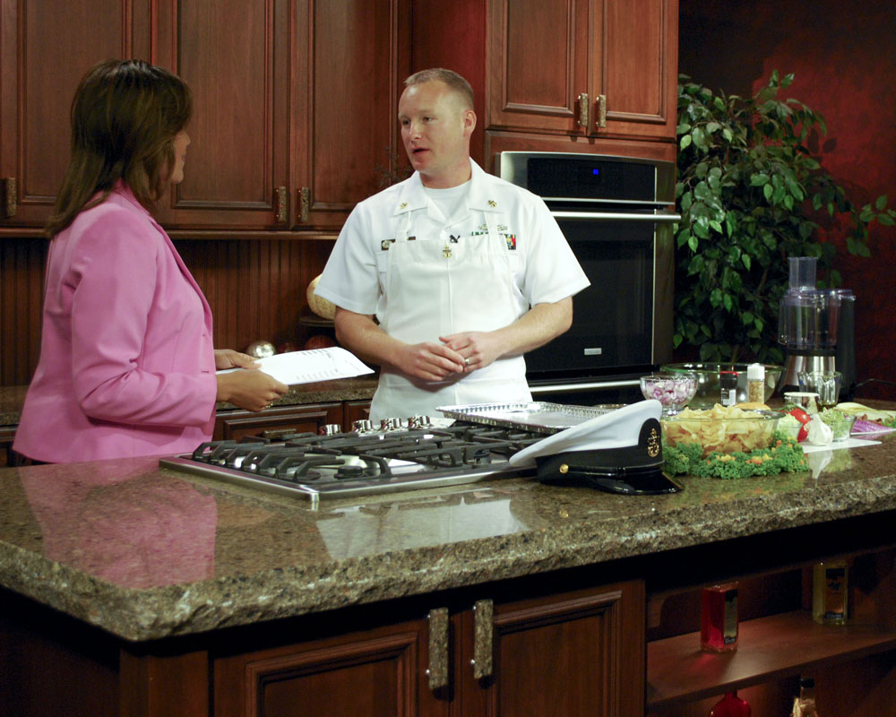 A cooking show host and guest behind a counter talking and preparing the recipe.