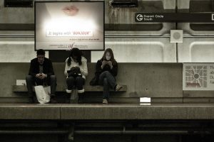 Three people sitting under a advert looking at their phone while waiting for a train.